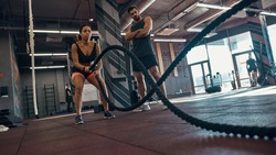 Bottom view of trainer looking at black girl doing exercise with sports rope in gym. Healthy lifestyle. Young concentrated woman and serious caucasian man wear sportswear. Fitness center in sunny day