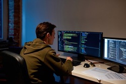 Concentrated male IT developer watching on computer monitor while programming script or code for mining cryptocurrency at home. Blockchain. Database. Modern digital payment and e-commerce. Night time
