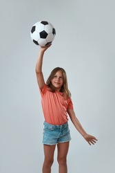 Front view of smiling little girl holding and playing with football ball. Caucasian female child wearing pink t-shirt and jean shorts. Childhood concept. White background. Studio shoot. Copy space