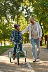 Caucasian father walk and help teenage son with cerebral palsy ride tricycle on pavement in green park. Family relationship and spend time together. Disability care and rehabilitation. Warm sunny day
