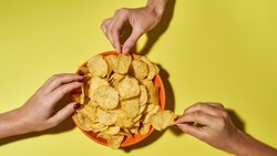 Partial female hands taking tasty and appetizing potato chips from bowl. Unhealthy eating and fast food. Crunchy snack for leisure. Isolated on yellow background. Studio shoot. Copy space. Top view