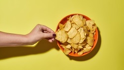 Cropped female hand taking delicious and appetizing potato chip from bowl. Unhealthy eating and junk food. Crispy snack for leisure. Isolated on yellow background. Studio shoot. Copy space. Top view