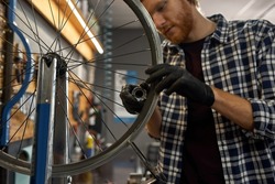 Young caucasian cycling repairman checking bicycle wheel spoke with bike spoke wrench in modern workshop. Bike service, repair and upgrade. Garage interior with tools and equipment