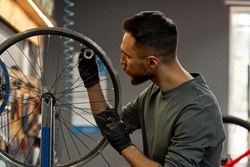 Side view of repairman checking bicycle wheel spoke with bike spoke wrench in modern workshop. Young caucasian bearded man. Bike service, repair and upgrade. Garage interior with tools and equipment