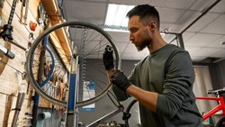 Young focused caucasian cycling technician checking bicycle wheel spoke with bike spoke wrench in modern workshop. Bike service, repair and upgrade. Garage interior with tools in sunny day