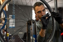 Cycling repairman checking bicycle wheel spoke with bike spoke wrench with blurred colleague on background in bicycle workshop. Young caucasian men. Bike service, repair and upgrade