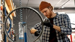 Cycling mechanic checking bicycle wheel spoke with bike spoke wrench in modern workshop. Young focused caucasian redhead man. Bike service, repair and upgrade. Garage interior with tools and equipment