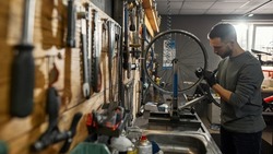 Side view of young caucasian cycling repairman checking bicycle wheel spoke with bike spoke wrench in modern workshop. Bike service, repair and upgrade. Garage interior with tools and equipment
