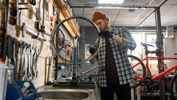Cycling repairman checking bicycle wheel spoke with bike spoke wrench in modern workshop. Young caucasian redhead man. Bike service, repair and upgrade. Garage interior with tools and equipment