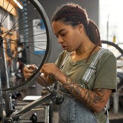 Young african american blonde female cycling master checking bicycle wheel spoke with bike spoke key in modern workshop. Bike service, repair and upgrade. Garage interior with tools and equipment
