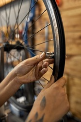 Partial image of female cycling repairman hands checking bicycle wheel spoke with bike spoke key in blured modern workshop. African american woman with tattoo. Bike service, repair and upgrade