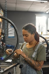 Young concentrated black female cycling repairman checking bicycle wheel spoke with bike spoke key in modern workshop. Bike service, repair and upgrade. Garage interior with tools and equipment