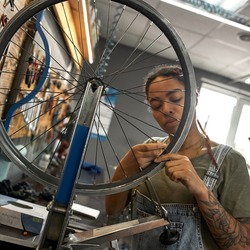 Young african american female cycling repairman checking bicycle wheel spoke with bike spoke key in modern workshop. Bike service, repair and upgrade. Garage interior with tools and equipment