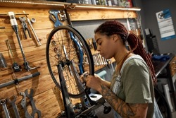 Young focused black female cycling master checking bicycle wheel spoke with bike spoke key in modern workshop. Bike service, repair and upgrade. Garage interior with tools and equipment