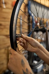 Cropped image of female cycling mechanic hands checking bicycle wheel spoke with bike spoke wrench in blured modern workshop. Black woman with tattoo. Bike service, repair and upgrade