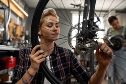 Young caucasian repairman girl installing bike cassette on wheel while her blurred black female colleague fixing electric scooter on background in modern workshop. Bike service, repair and upgrade