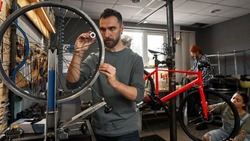 Male cycling repairman checking bicycle wheel spoke with bike spoke key while working with colleagues on background in workshop. Young multiracial men and women. Bike service, repair and upgrade