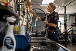 Side view of young european blonde female cycling technician checking bicycle wheel spoke with bike spoke wrench in workshop. Bike service, repair and upgrade. Garage interior with tools and equipment