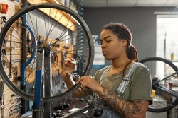 Young african american female cycling mechanic checking bicycle wheel spoke with bike spoke key in modern workshop. Bike service, repair and upgrade. Garage interior with tools and equipment