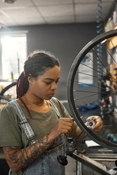 Young african american female cycling worker checking bicycle wheel spoke with bike spoke key in modern workshop. Bike service, repair and upgrade. Garage interior with tools and equipment