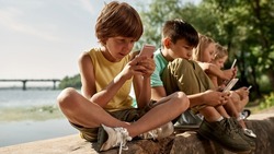 Row of multiethnic children using and browsing mobile phones on log on river coast outdoors. Boys and girl of generation alpha. Gadget addiction. Childhood lifestyle. Sunny summer day