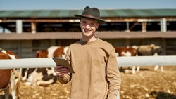 Front view of smiling male farmer with digital tablet near blurred paddock with milk cows on farm or ranch. Young caucasian guy looking at camera. Modern countryside lifestyle. Agriculture. Sunny day