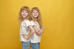 A portrait of twin girls. Two beautiful and positive curly blonde sisters who are 6 or 7 years old, wearing white t-shirts and casual jeans, standing back to back on a yellow background.