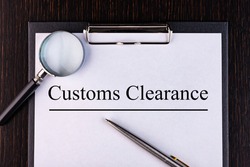 Text CUSTOMS CLEARANCE is written on a notebook with a pen and a magnifying glass lying on the table. Business concept.