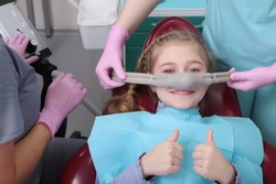 A little girl is comfortable to treat her teeth under superficial sedation. The girl smiles and holds two thumbs up. Milk teeth treatment. Treatment of children's teeth with nitrous oxide. 