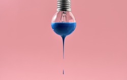 Filament bulb with blue paint on the pink background in the studio. Paint is dripping down from it. Closeup. Horizontal.