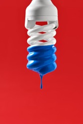Spiral fluorescent bulb with blue paint on the red background in the studio. Paint is dripping down from it. Closeup. Vertical.