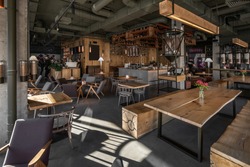 Restaurant in a loft style with textured wooden walls and concrete columns. There are tables with chairs, many shelves with bottles, glass coffee bean dispensers, coffee machines and equipment.