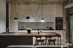 Kitchen in a loft style with concrete and brick walls. There is a kitchen island with a sink, plant and black chairs, light lockers with built-in oven and fridge, dark tabletop with stove and teapot.