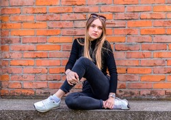 young beautiful girl 17 years old with long hair in a black sweater and jeans, sits against the background of a brick wall