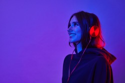 Fashion hipster girl smiles and wear headphones listening to music over color neon background at studio.