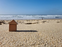 Small miniature kids wooden toy home model on sunny sand beach with blue sky and white clouds and ocean background. Copy space of family lifestyle and business real estate property investment concept