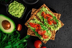 Avocado sandwich with avocado cream and rye crisp bread for snack. Fiber, fitness and diet food. Rye bread with guacamole, arugula and cherry tomatoes on dark background. High quality photo
