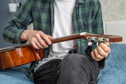 A man in a green plaid shirt tunes a guitar. A musician tunes a classical guitar while sitting on a bed during quarantine. The concept of self-isolation, online learning.