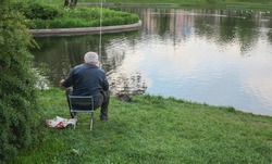 An elderly male fisherman sits with a fishing rod on the shore of a lake in a city park. An elderly man is fishing in a city park in the evening twilight. The concept of active recreation.