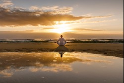 yoga meditation silhouette lotus sunrise beach, mindfulness, wellness and wellbeing concept, water reflection of man in yoga lotus pose sitting alone on sand with ocean cloud background, copy space