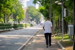 Back view of a young man wearing a white t-shirt and long black trousers walking on the footpath.