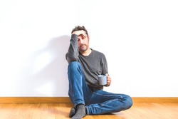 attractive man on his 40s dressing casual sitting on the wooden floor of an apartment with gesture of tiredness or concern with white wall as background