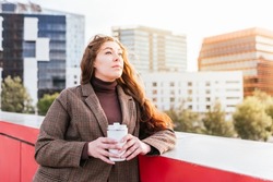 Pensive female in outerwear with long hair. She is looking away and thinking while leaning on bridge border and enjoying takeaway coffee on sunny day in city