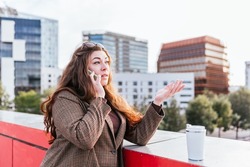 Woman in outerwear with long hair looking away and answering phone call. while leaning on bridge border near zero waste cup of coffee to go on blurred background of city city