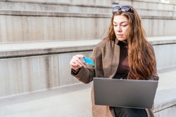 Female in outerwear with long hair using credit card and laptop to make online purchase while sitting on steps on city street
