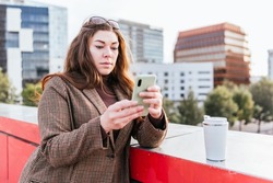 Woman in outerwear with long hair browsing social media on mobile phone. She is leaning on bridge border near eco friendly cup of hot drink in modern city
