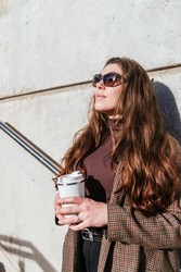 Stylish woman in outerwear with sunglasses leaning on wall and looking away while enjoying coffee to go and dreaming on city street