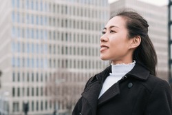 Happy Asian businesswoman in smart casual outerwear looking away with smile and admiring city street on blurred background of high rise buildings