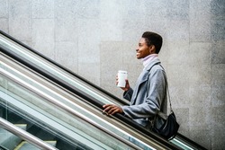 Side view of stylish African American female in stylish coat and sweater. She is with cup of coffee and bag smiling and riding moving stairs in city