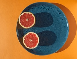 Half pink sliced grapefruit on blue plate on orange background. Citrus pink fruit. Modern food trendy concept.  Food still life. Vitamin C, antioxidant. Blue hand made ceramic. Red juicy fruit.